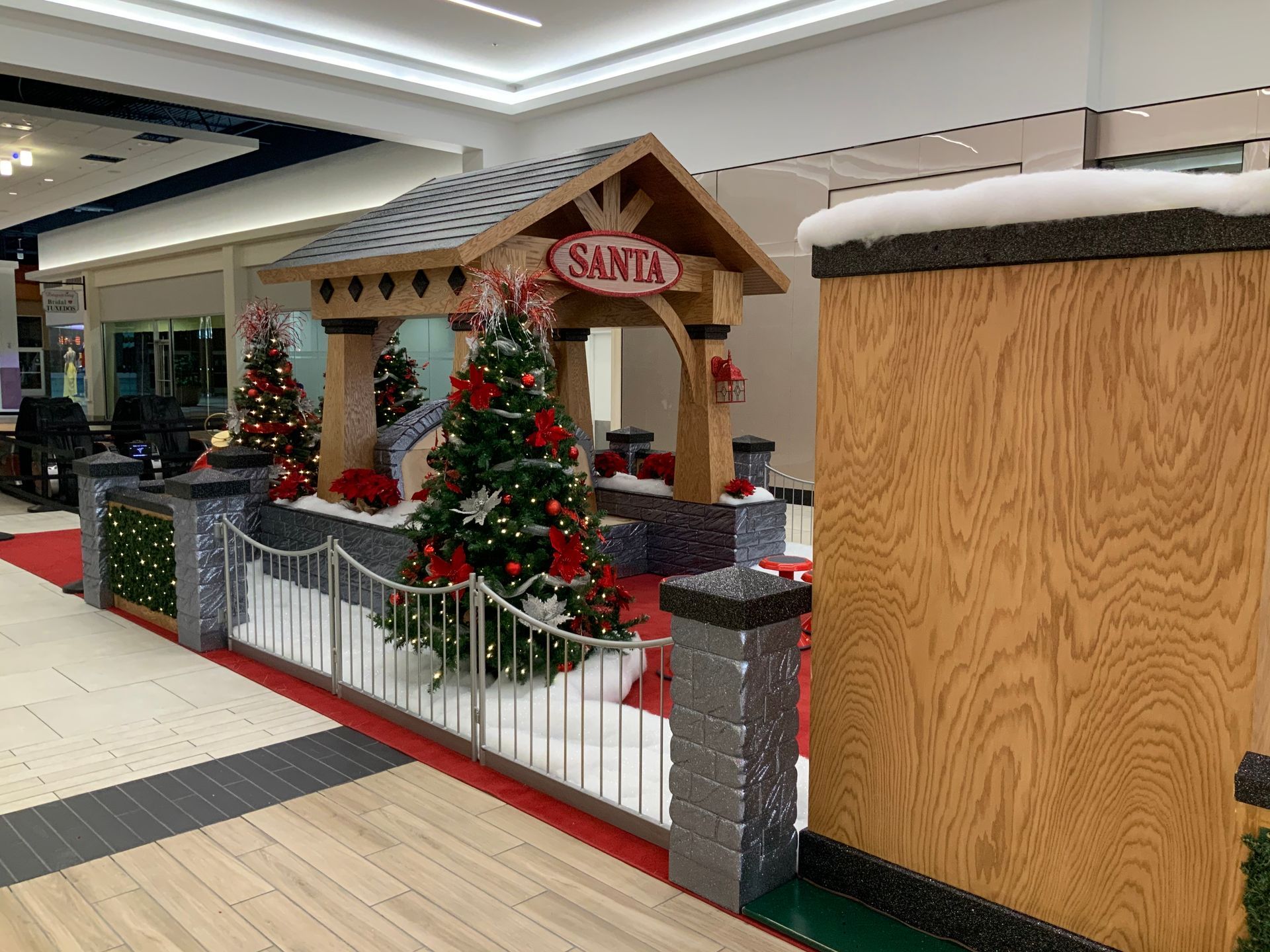 Santa's display in a mall with a wooden house, Christmas trees, and a fence, on a red carpet.