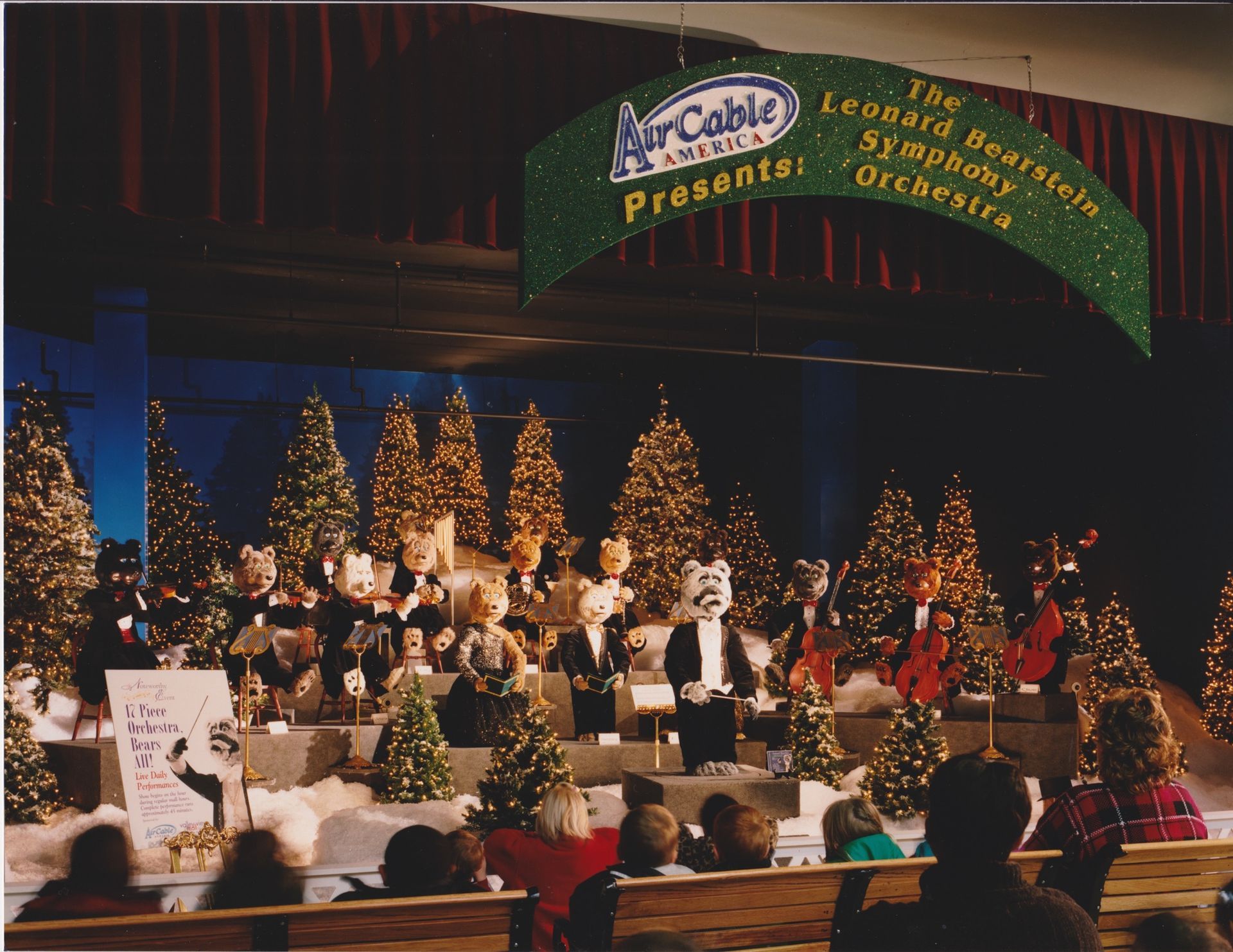Orchestra of cats in formal wear performing on a stage with Christmas trees, beneath a banner.