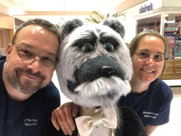 Two adults flank a large, fuzzy bear puppet in a mall. They smile for the camera, all wearing matching shirts.