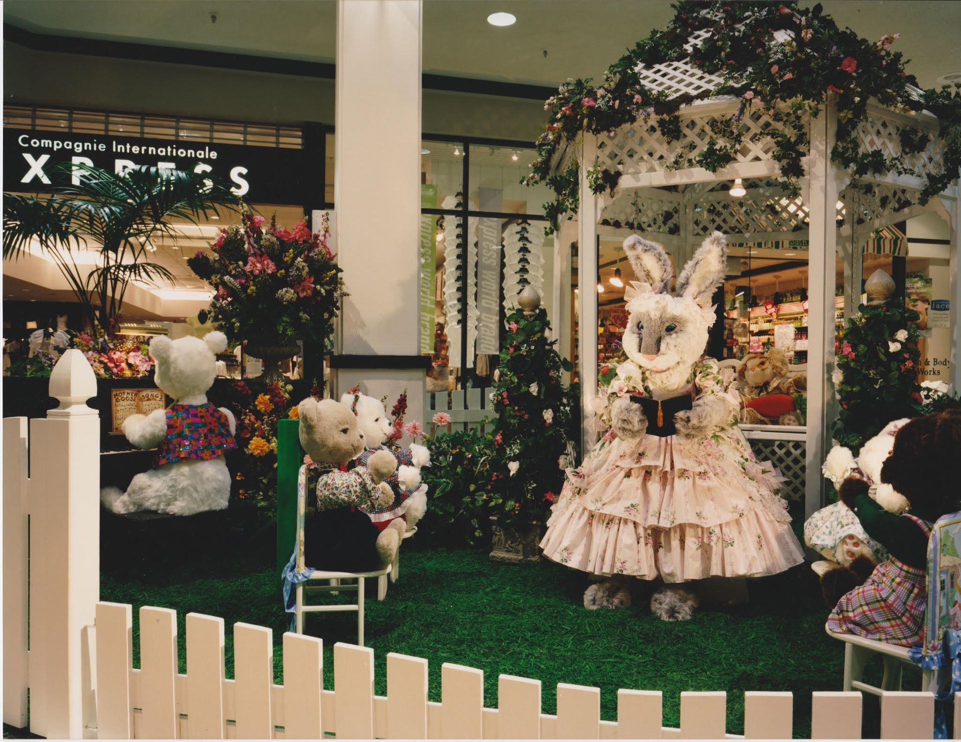Easter display in a mall with a rabbit in a pink dress and teddy bears near a white fence.