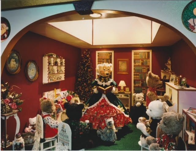 A Christmas-themed room with teddy bears. A queen bear reads while others listen. Red walls, a tree, and gifts.