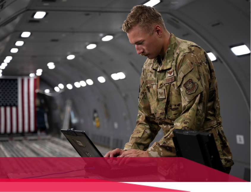 Airman in uniform works on a laptop inside a cargo plane. American flag visible.