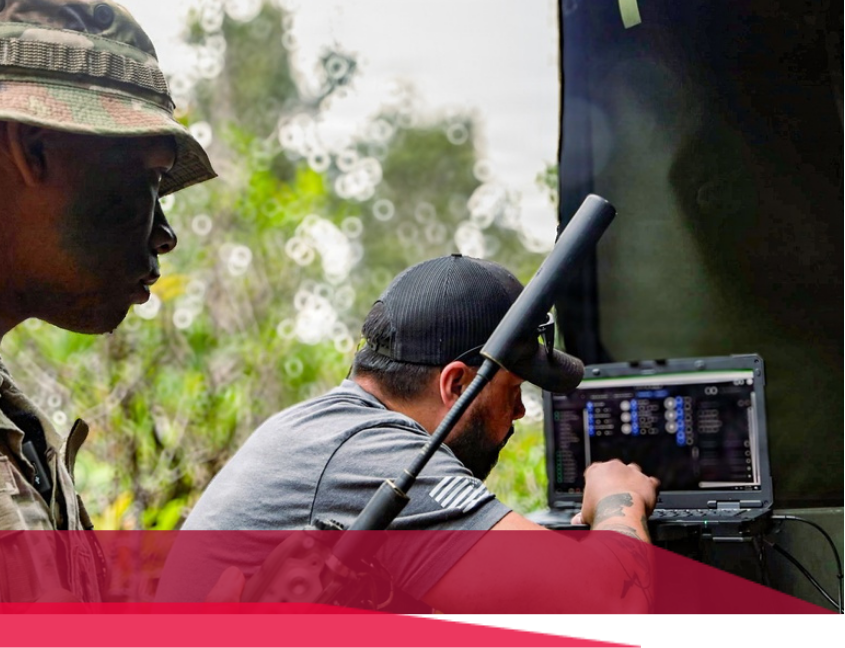 Two people operating a laptop outdoors. One wears a camouflage hat. One has a tattoo and is focused on the screen.