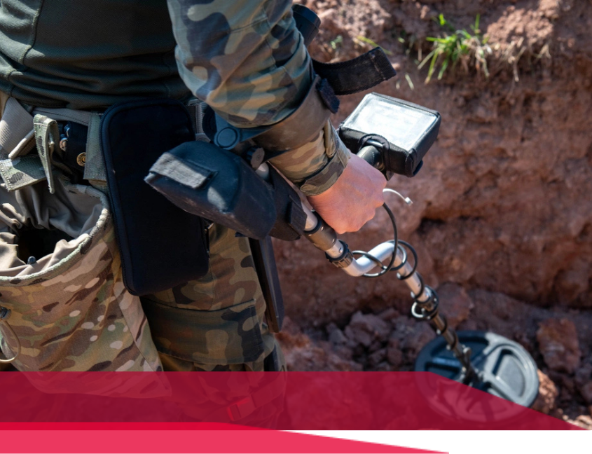 Person in camouflage using a metal detector outdoors near a dirt wall.
