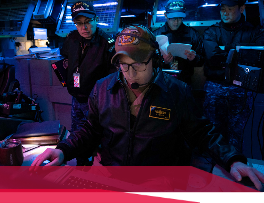 Navy personnel at computer consoles in a dimly lit control room, focused on tasks.