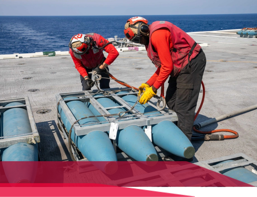 Two people in red vests loading torpedoes on a naval vessel's deck, near the sea.