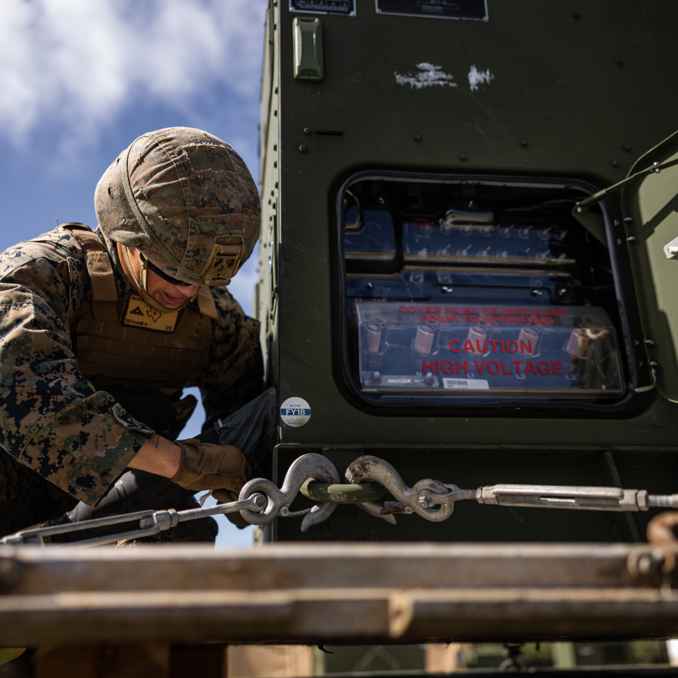 A man in a camouflage uniform is standing in front of two computer monitors.