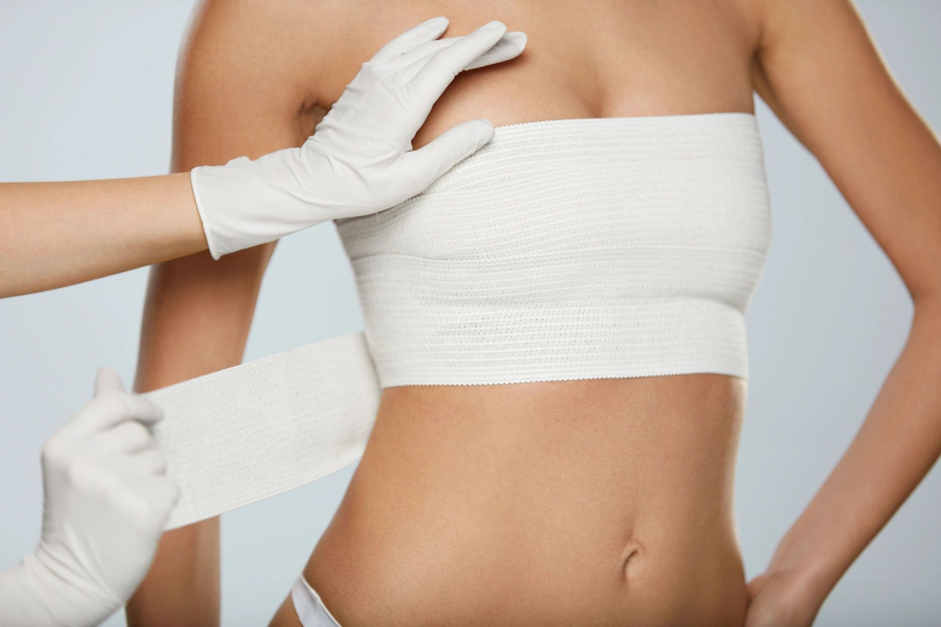 Person's torso bandaged after surgery, hands in gloves placing dressing. Light skin, pale blue background.
