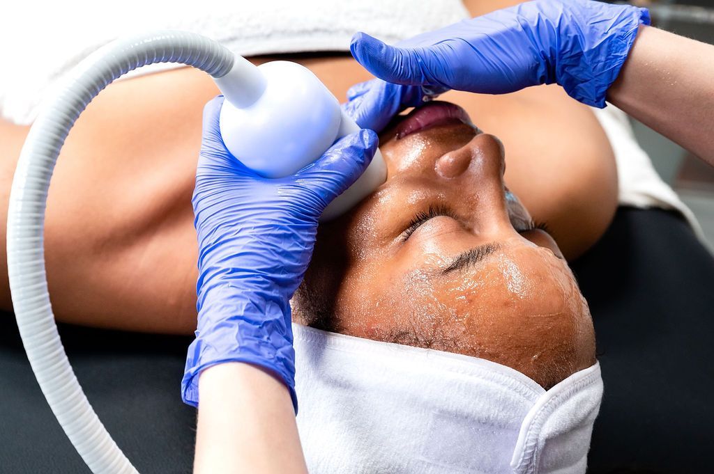 Person receiving facial treatment with a device held by gloved hands; towels and hose visible.