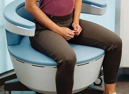 Woman seated on a light blue and white medical chair with arms, wearing a maroon top and brown pants.