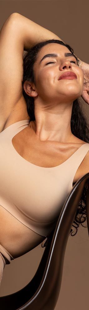 Woman in beige top smiles while leaning back in a chair, arm raised behind her head, lit by soft lighting.
