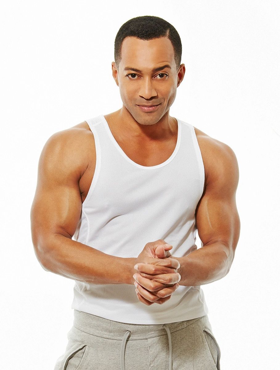 Man in white tank top and gray sweatpants, smiling with hands clasped, against white background.