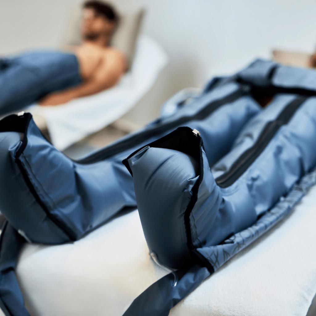 Person in blue compression boots, resting on a massage table. Another person is in the background.