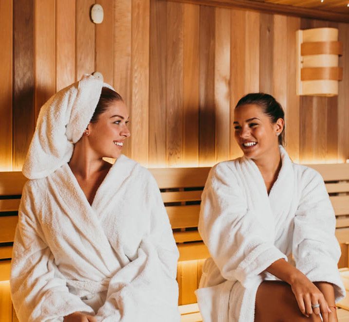 Two women in white robes smiling, seated in a wooden sauna.
