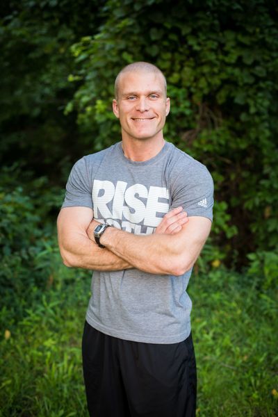 A man in a gray shirt with the word rise on it is standing in the grass with his arms crossed.