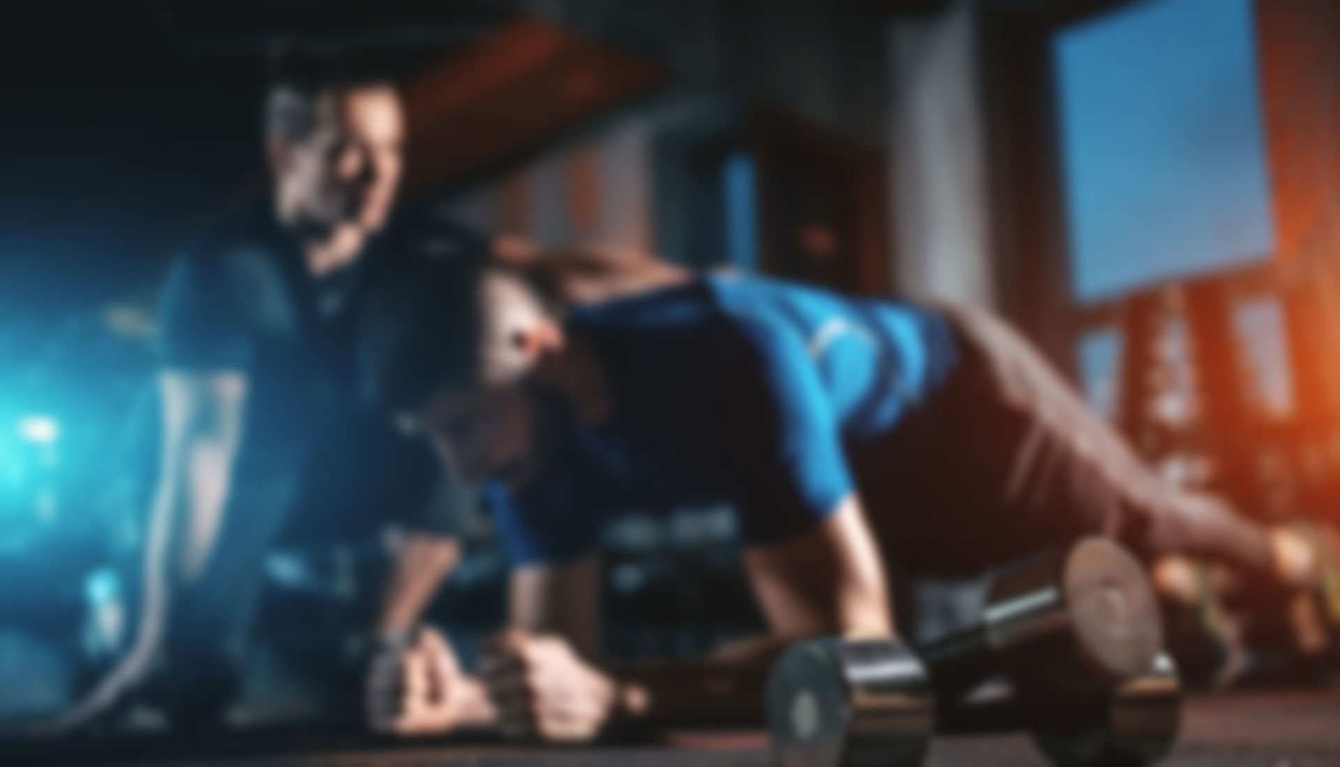 A person performs a plank exercise on a gym floor with a trainer observing nearby, a dumbbell placed in the foreground.