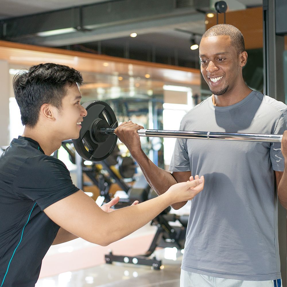 A man is helping another man lift a barbell in a gym.