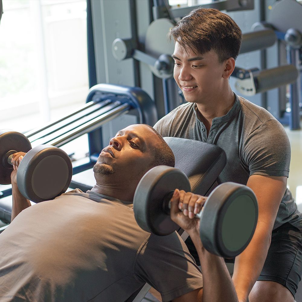 A fitness trainer spots a man performing dumbbell chest presses on a weight bench in a gym.
