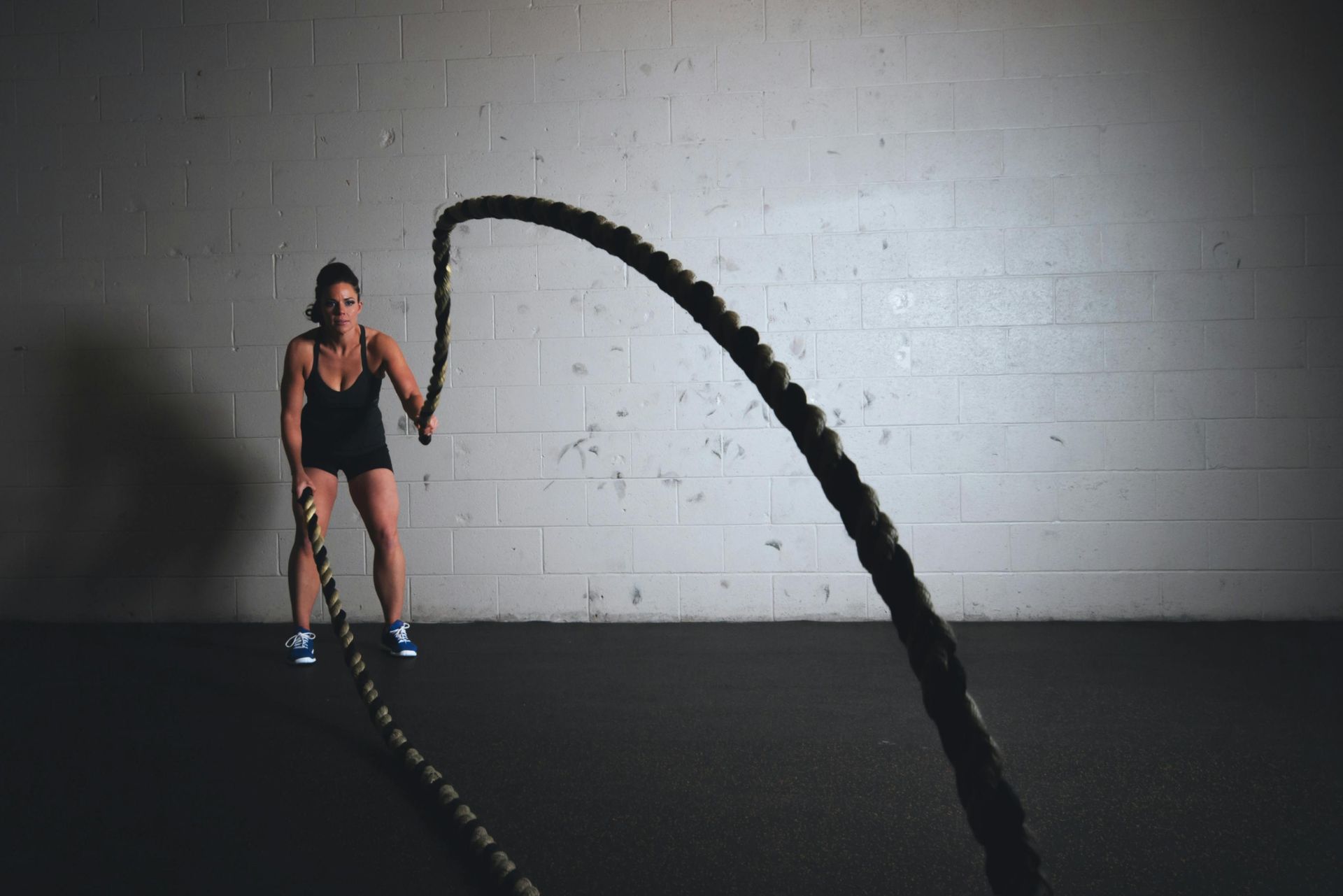 Woman using battle ropes in a gym; dark setting, black ropes, intense workout.