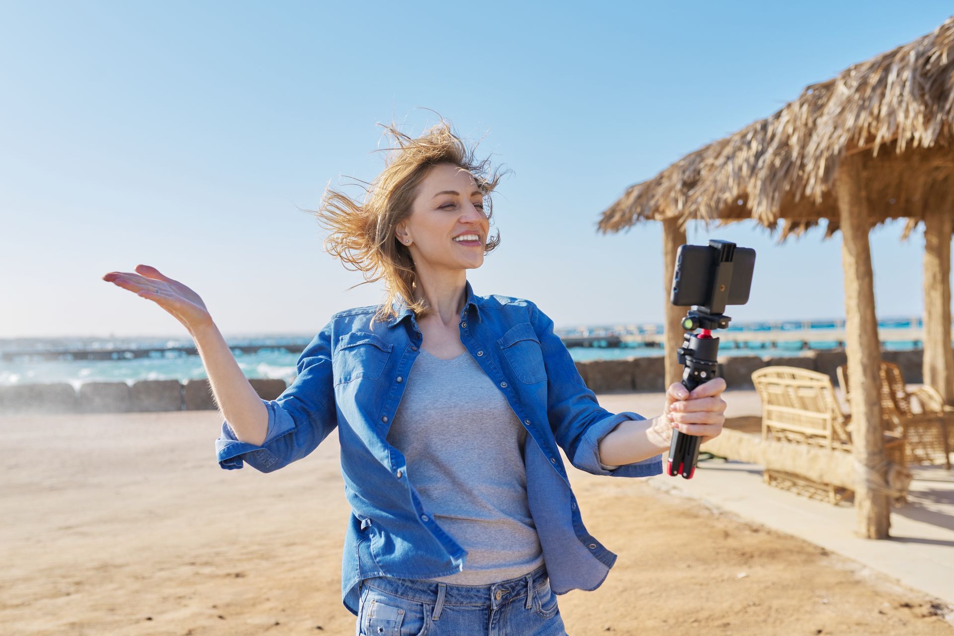 Woman vlogging with phone on beach, windblown hair, smiling, holding out hand. Blue sky, thatched shelter.