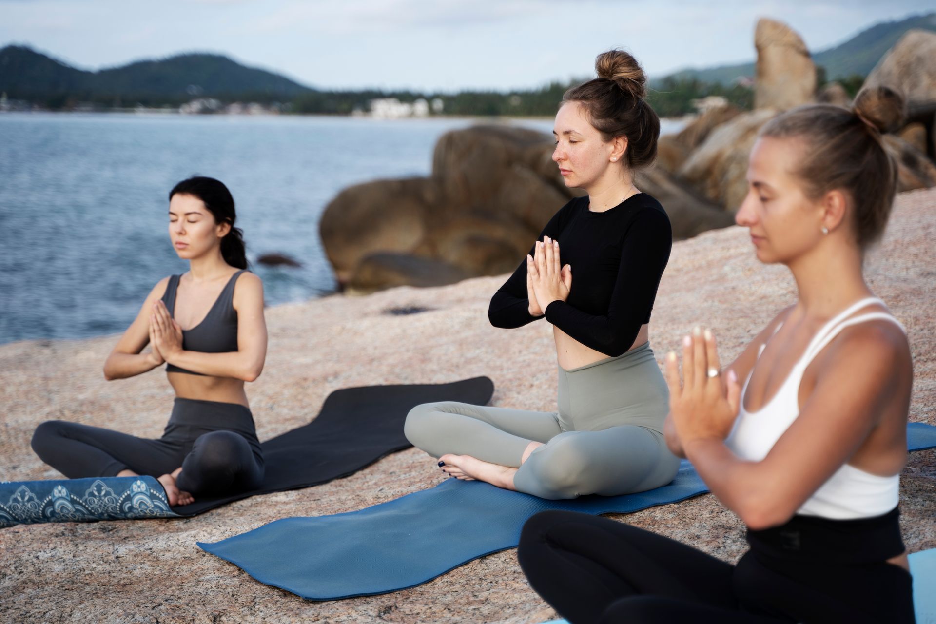 Three people meditating with hands in prayer pose on yoga mats near a rocky beach.