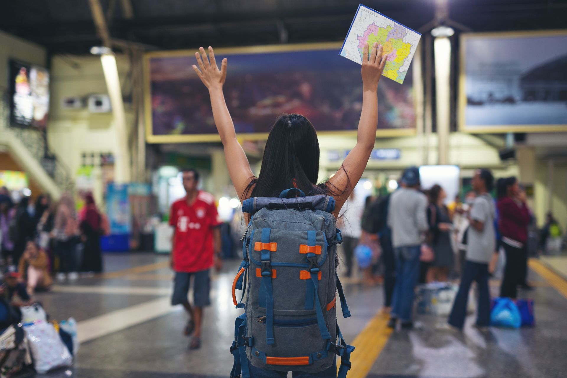 Woman with backpack raises arms, holding a map in a bustling train station.