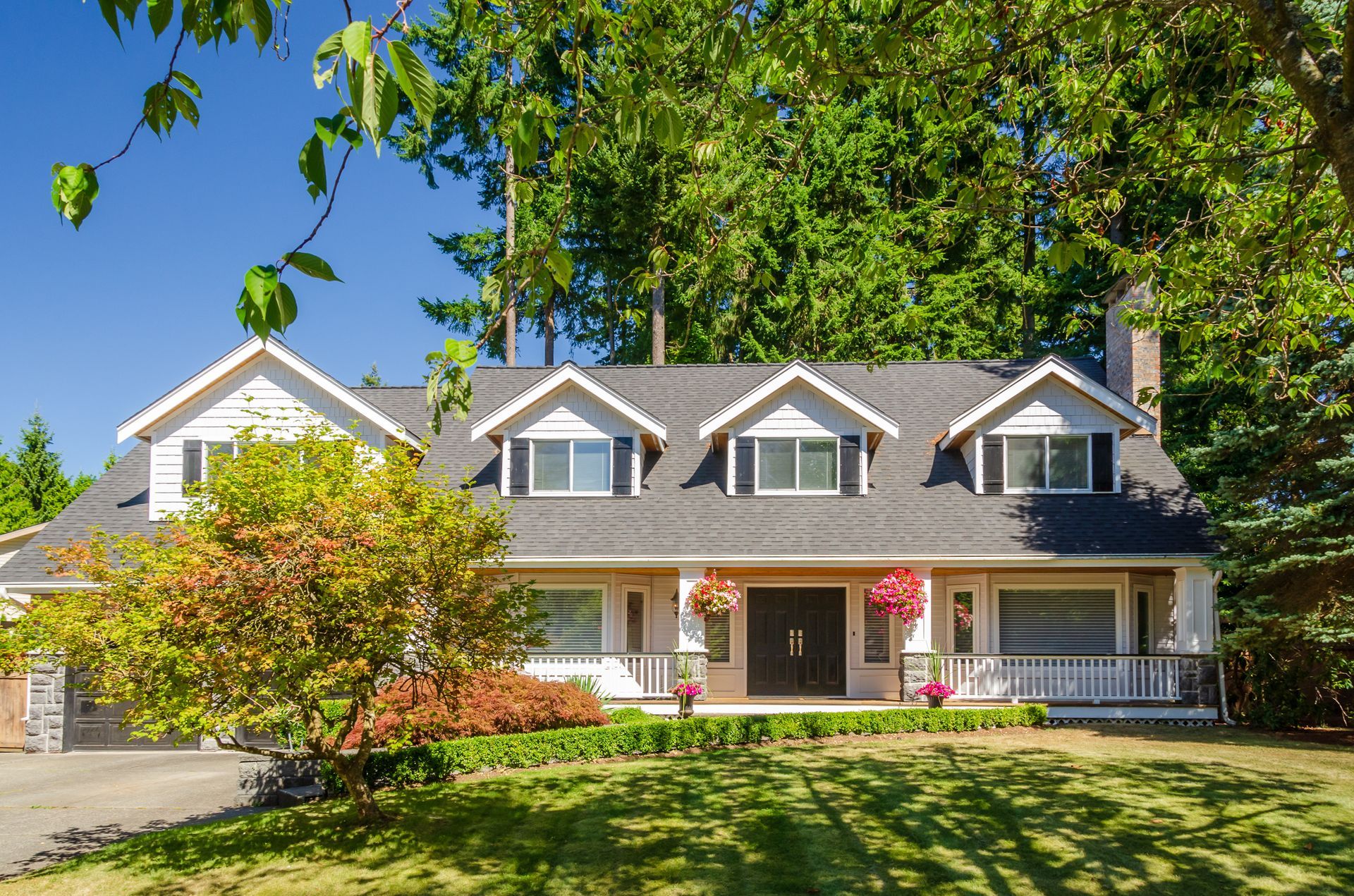 White house with dark gray roof, dormers, and front porch, surrounded by trees and green lawn.