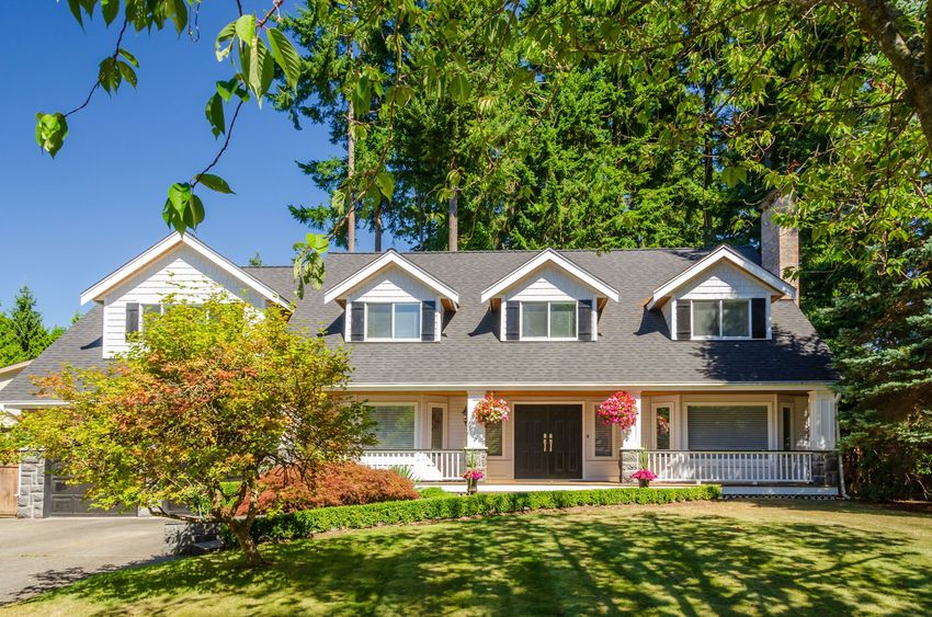 White house with dark gray roof, dormers, and front porch, surrounded by trees and green lawn.