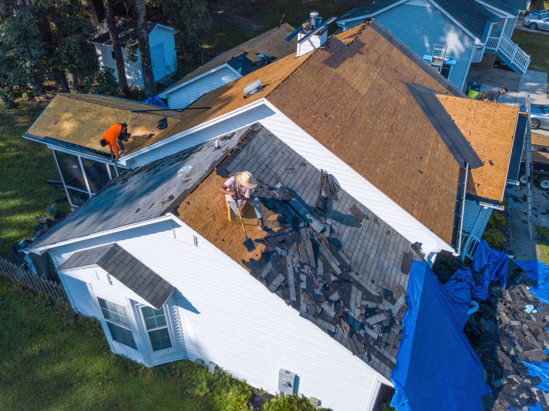 Aerial view of house roof being repaired; workers on roof; blue tarps; brown and grey shingles.