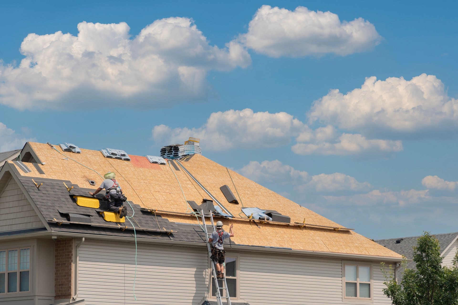 Roofers installing shingles on a house roof under a blue sky with fluffy clouds.