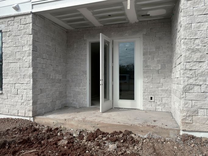 Vista esterna: muri in pietra grigio chiaro, patio in cemento, doppie porte aperte, terra in primo piano.
