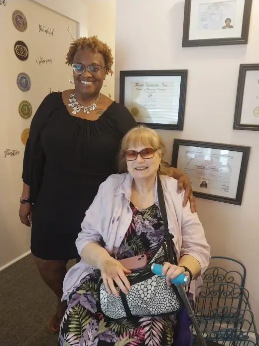 Two women posing in an office, one standing behind a seated woman holding a cane.
