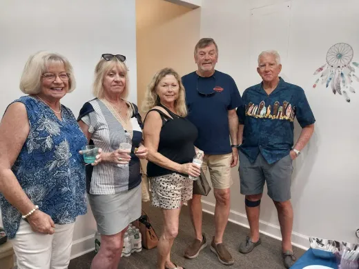Five people posing indoors in casual clothes, smiling beside a wall with a decorative dreamcatcher.