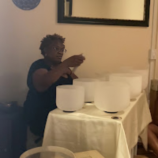Person seated at a table with several white singing bowls, reaching toward one bowl in a dim room.