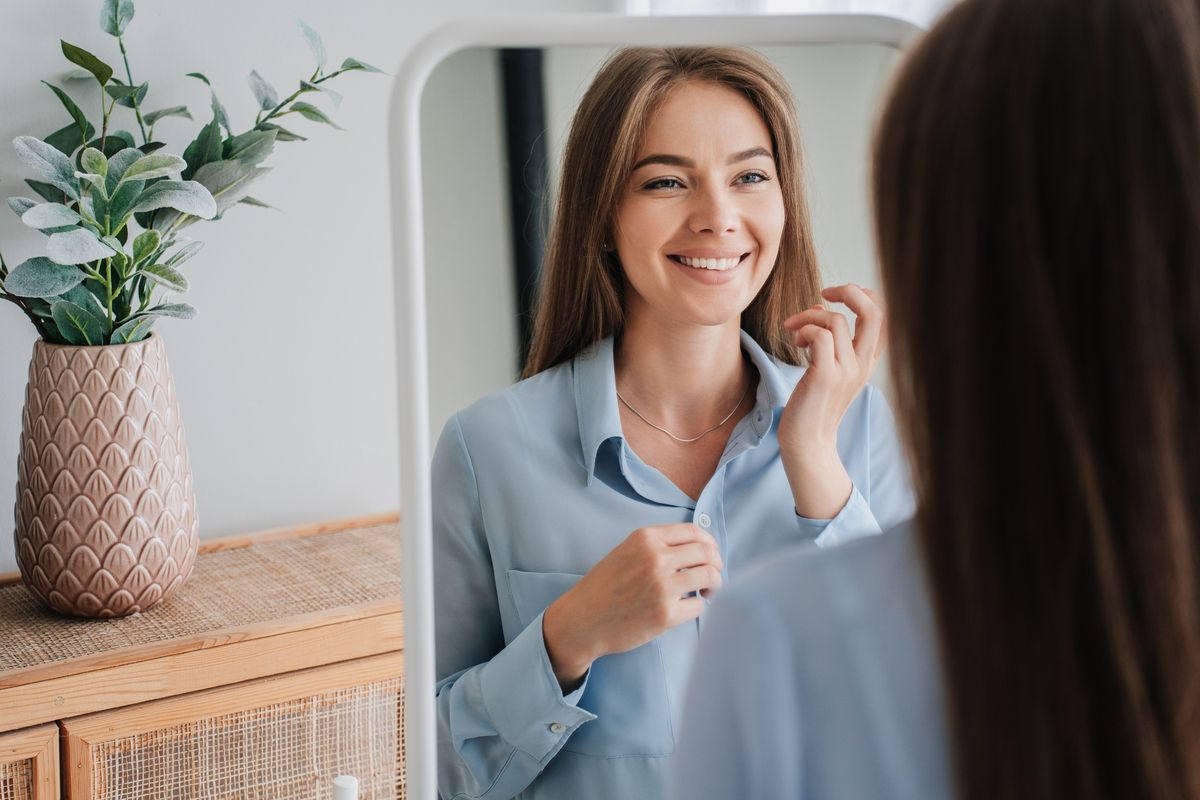 Smiling woman in a light blue blouse adjusting her collar in a mirror beside a vase of flowers
