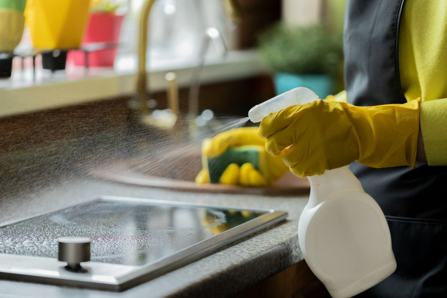 A person wearing yellow rubber gloves sprays cleaner onto a kitchen stovetop while holding a sponge in their other hand.
