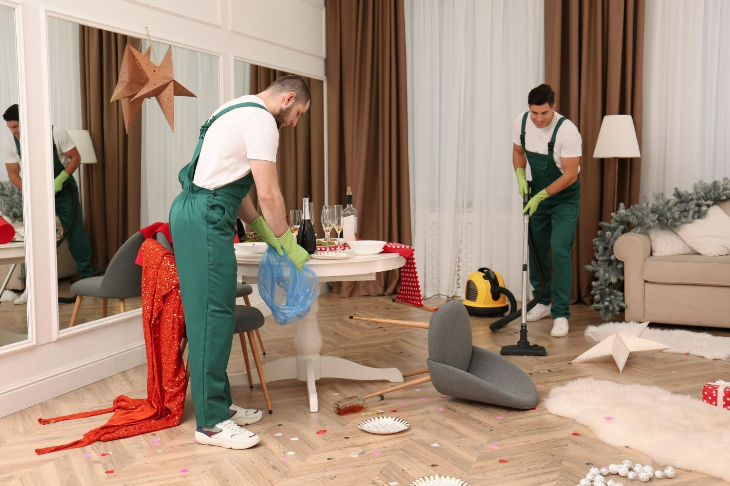 Two cleaners in green overalls tidy a living room scattered with holiday decorations after a party.