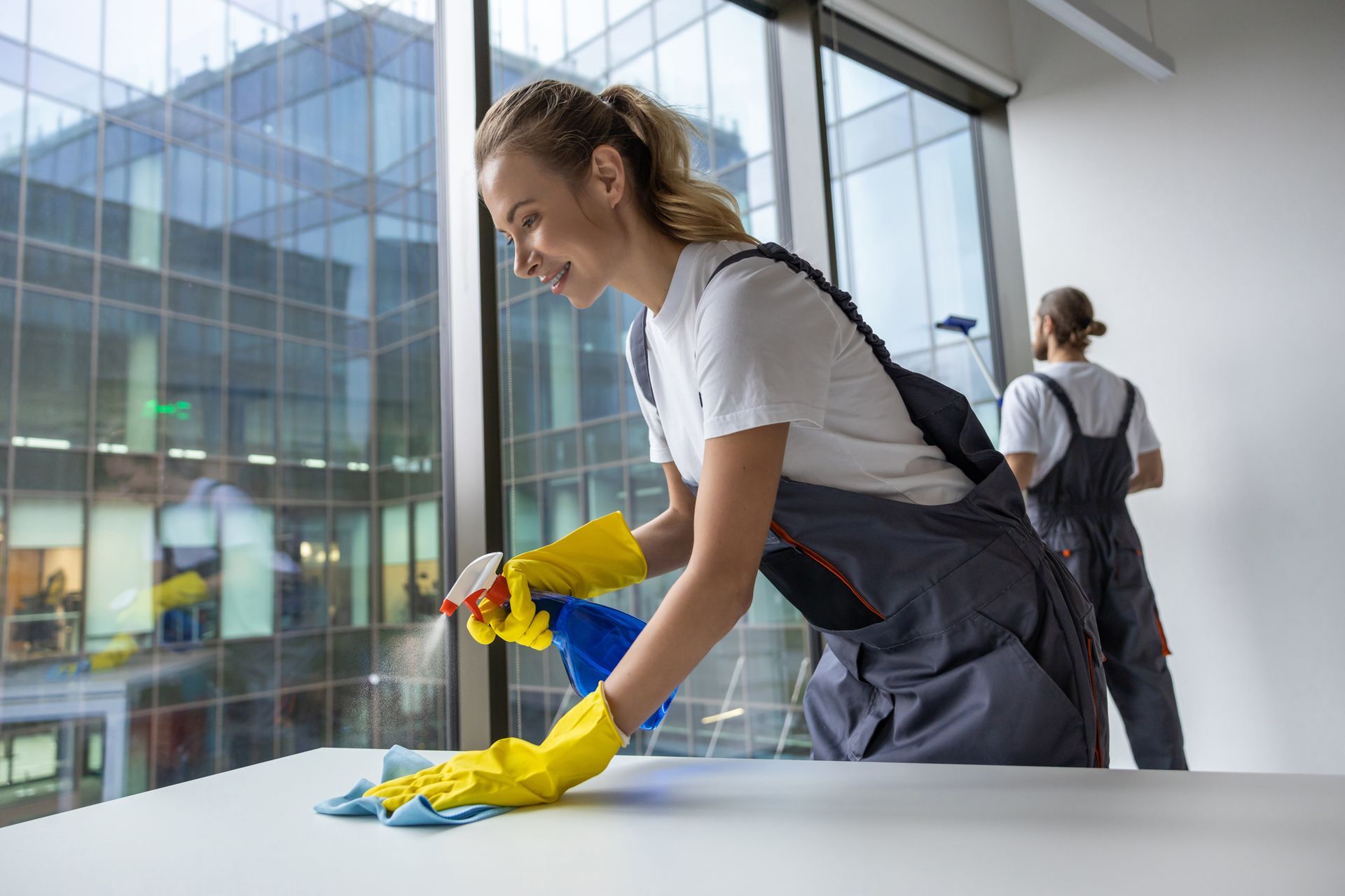 A cleaner in grey workwear and yellow gloves wipes a white surface with a spray bottle, with a colleague working nearby.