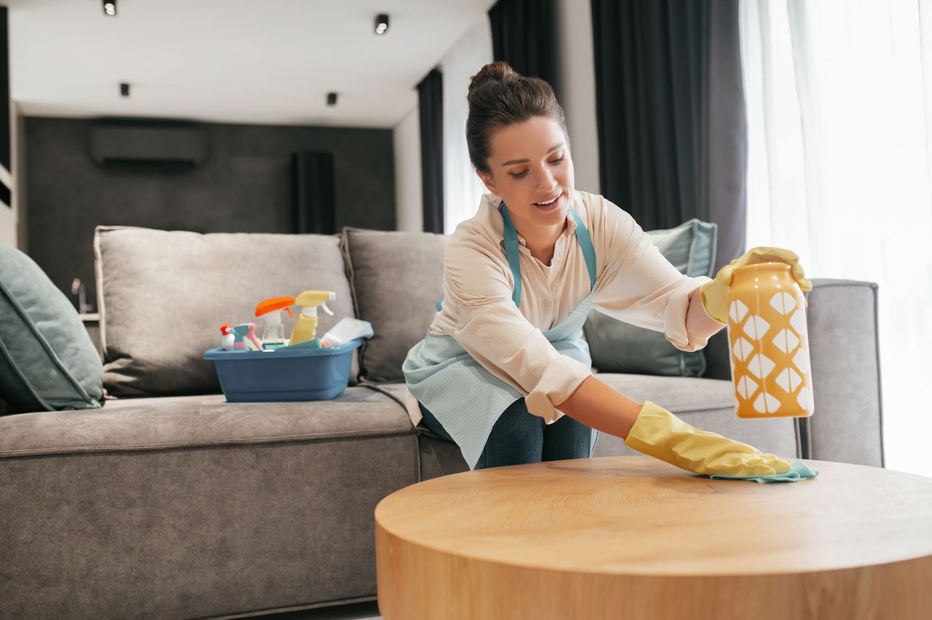 Woman in gloves dusting a wooden coffee table in a living room.