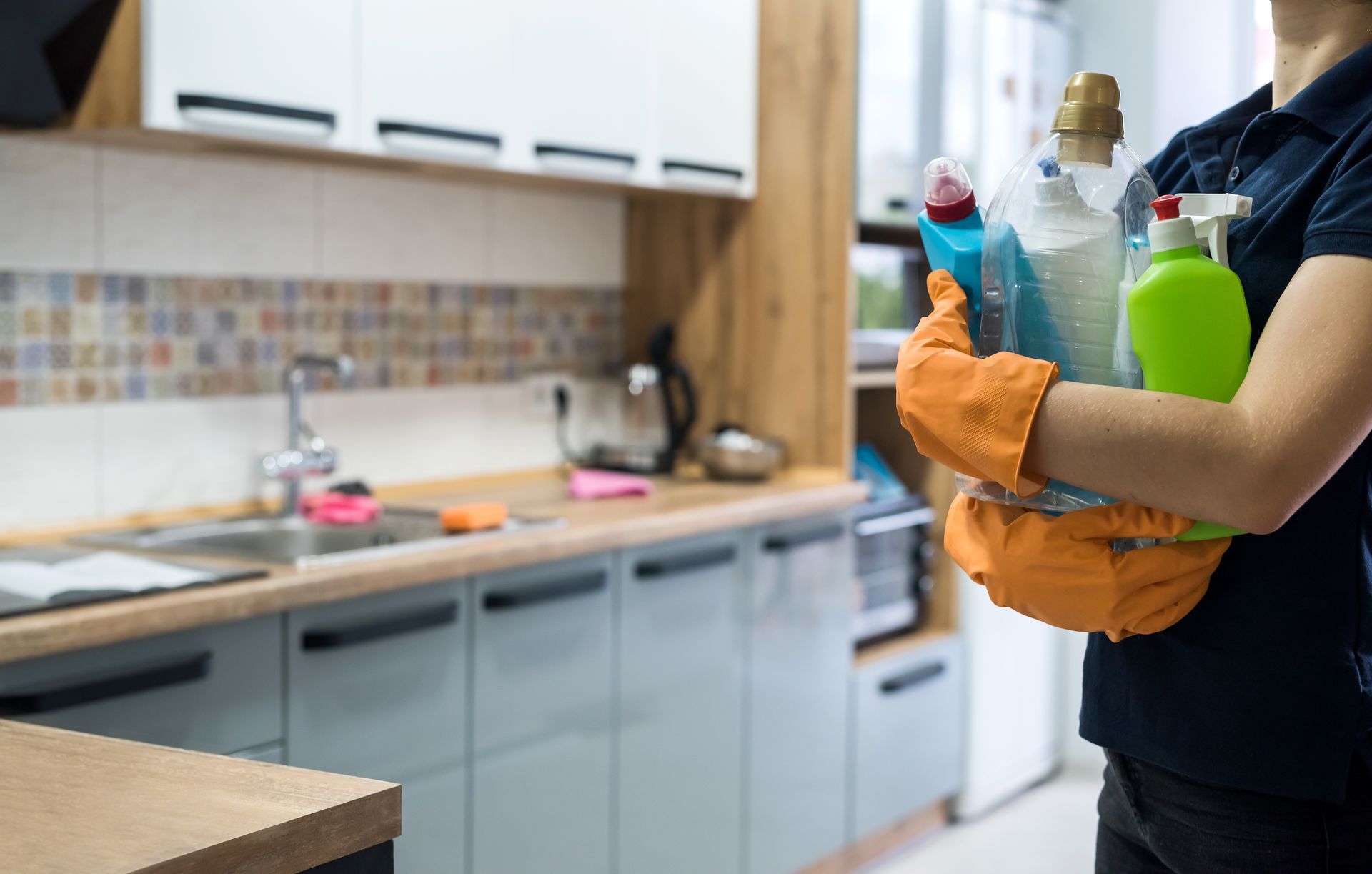Person wearing orange gloves holding cleaning supplies in a kitchen.