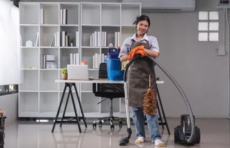 Woman in apron leans on vacuum cleaner in office, smiling.