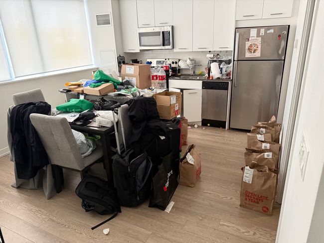 Cluttered kitchen and dining area with boxes, bags, and items piled on chairs and counters