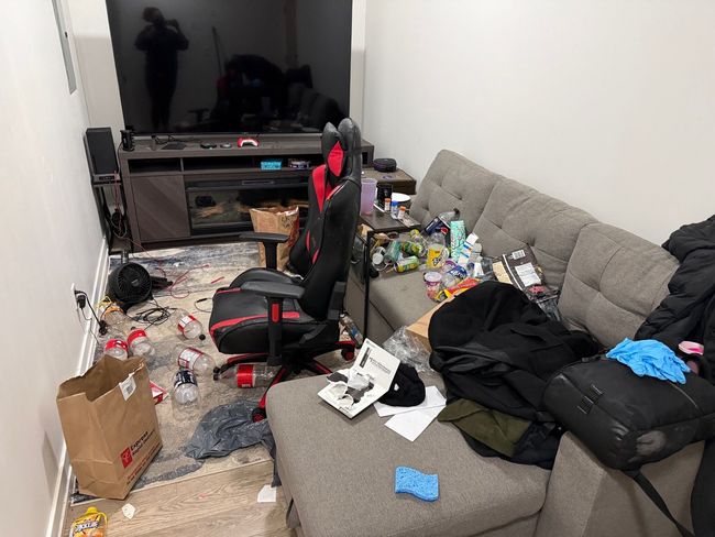 Cluttered living room with sofa, gaming chair, TV, scattered items, and trash on the floor