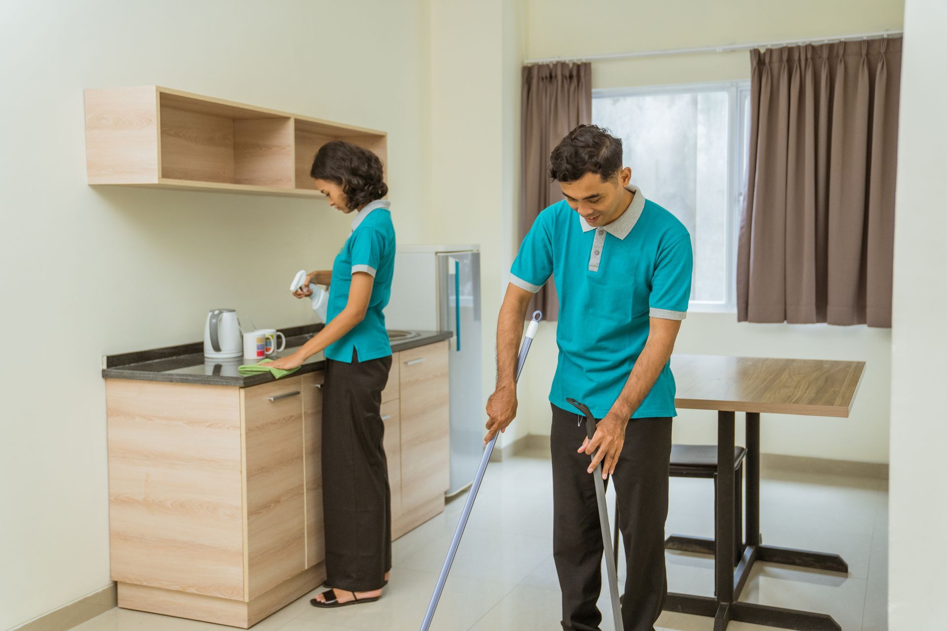 Two people in matching teal shirts and dark pants cleaning a kitchen; one wipes a counter while the other mops the floor.