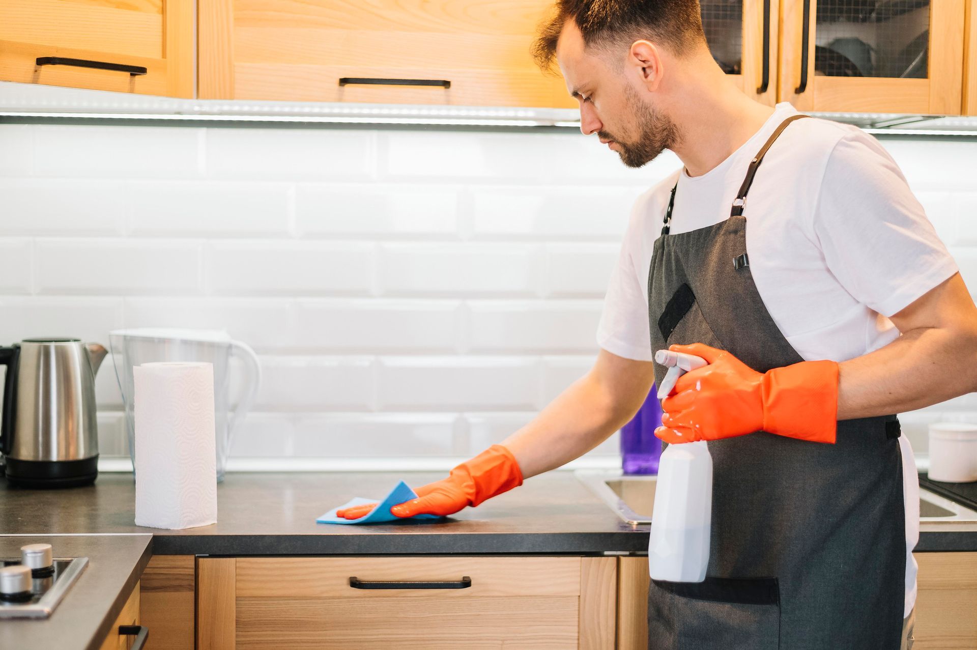 A man is cleaning a kitchen counter with a cloth and spray bottle.