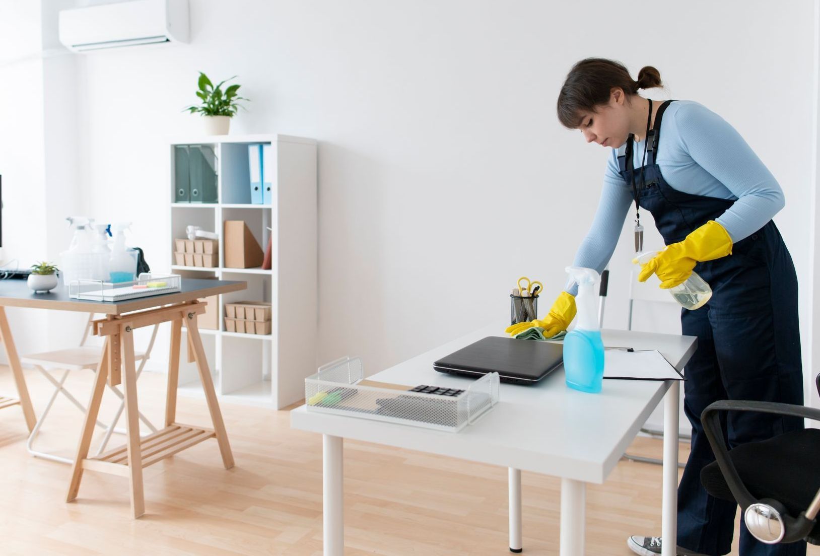 Woman in blue jumpsuit and yellow gloves cleaning an office desk.