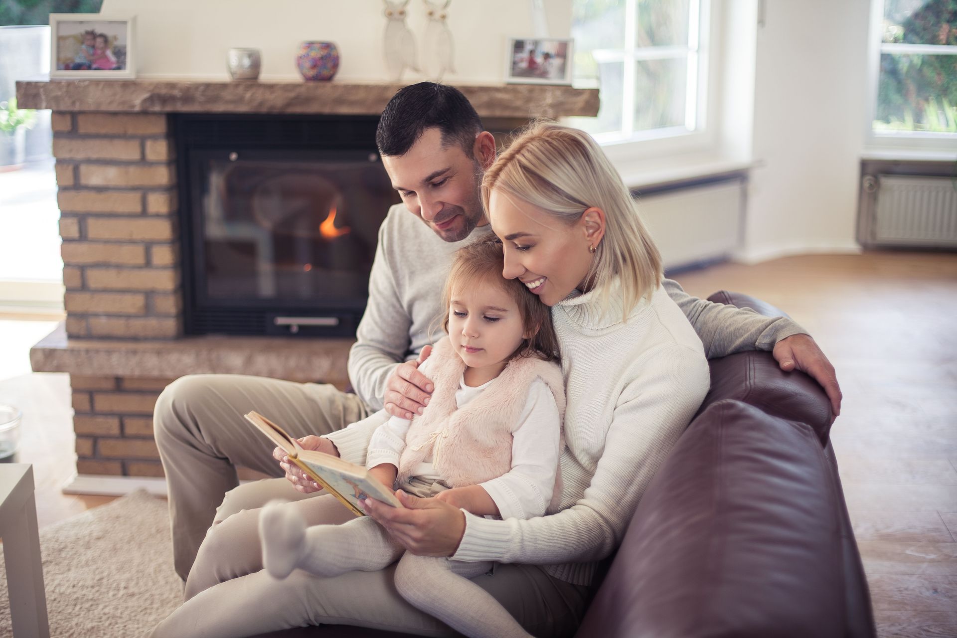 A family is sitting on a couch reading a book in front of a fireplace.