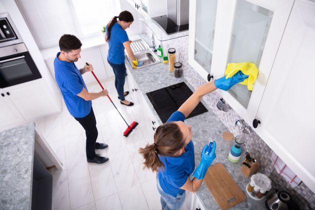 Three people cleaning a bright kitchen countertop and sink with mops, cloths, and spray bottles