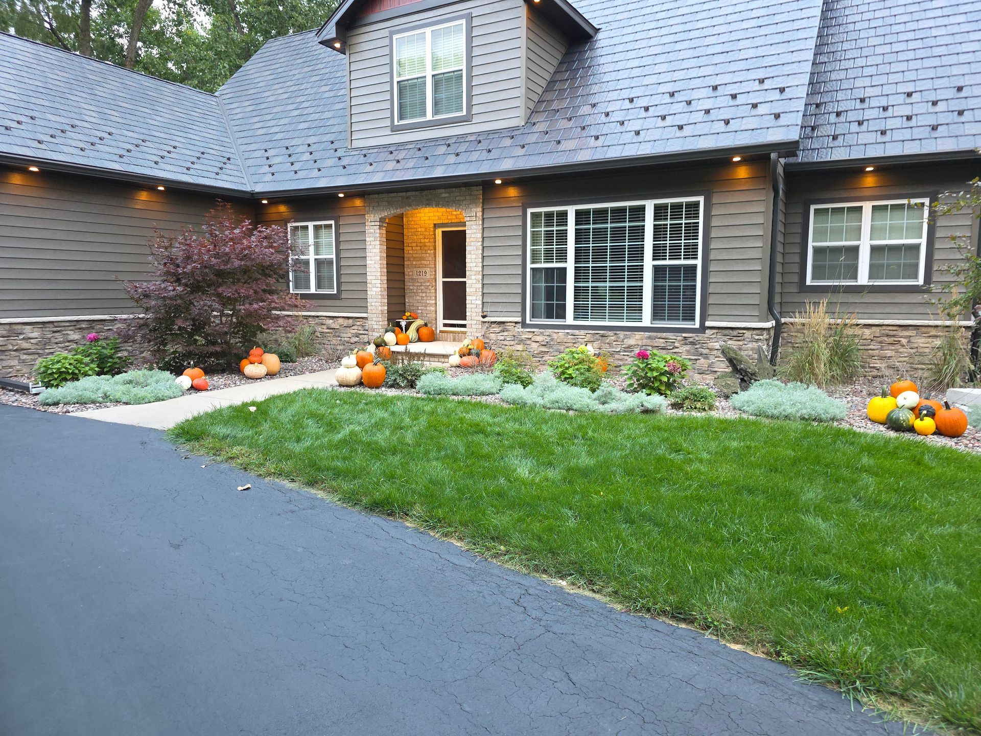 House with pumpkins, green lawn, dark asphalt driveway, and gray siding.