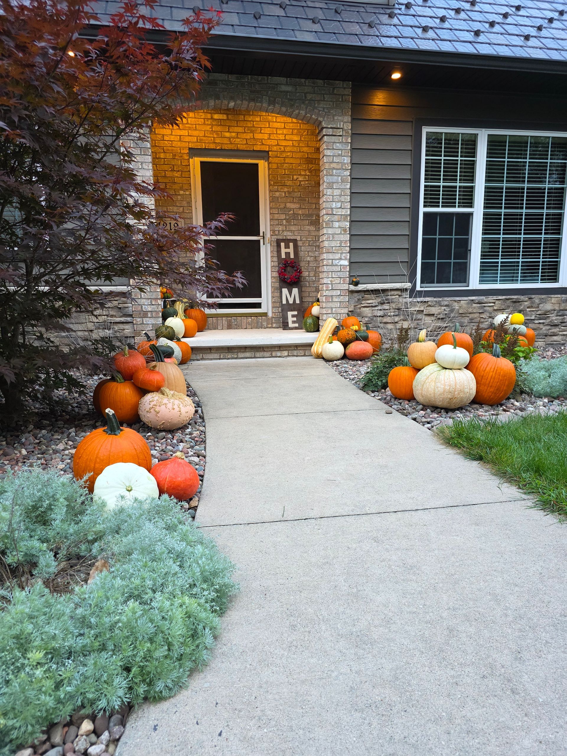 A walkway lined with pumpkins leads to a front door decorated with autumn decor; a house in the background.